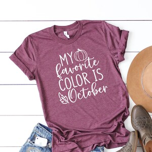 May include: A maroon t-shirt with the text "MY favorite COLOR IS October" in white, along with a pumpkin and leaf graphic. The shirt is paired with denim shorts, brown leather boots, and a tan hat.