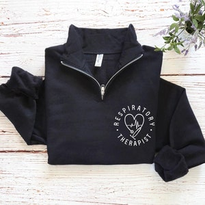 May include: Black quarter-zip sweatshirt with white text and a heart-shaped design. The design includes a stethoscope and the words "Respiratory Therapist" in a circular pattern. The sweatshirt has a high collar and long sleeves.