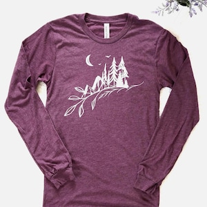 May include: A long-sleeved, burgundy t-shirt featuring a white graphic of a mountain scene with trees, a crescent moon, and a branch with leaves. The shirt has a crew neck and is made of a soft material.