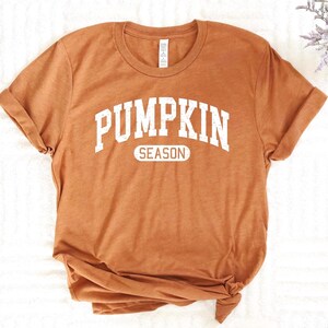 May include: A burnt orange t-shirt with rolled-up sleeves. The shirt features the text "PUMPKIN SEASON" in a distressed white font, perfect for autumn. The shirt is knotted at the bottom.