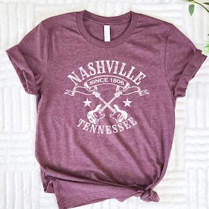 May include: Maroon t-shirt with a white graphic that reads "Nashville Since 1806 Tennessee" with crossed guitars and stars. The shirt is made of a soft material and has rolled-up sleeves.