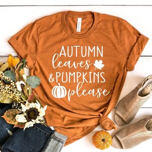 May include: An orange t-shirt with the text "AUTUMN leaves & PUMPKINS please" in white. The shirt is styled with a knot at the bottom. The image includes a pair of jeans, sandals, and fall-themed decorative items.