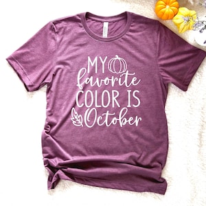 May include: A mauve short-sleeved t-shirt with the text "MY favorite COLOR IS October" in white script. The design includes a pumpkin and a leaf. The shirt is a casual fall-themed apparel item.