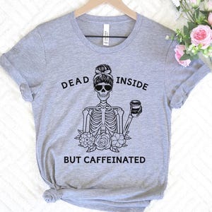 May include: A gray t-shirt with the words "DEAD INSIDE BUT CAFFEINATED" printed in black. The graphic features a skeleton wearing sunglasses and holding a coffee cup. Floral accents are at the bottom of the skeleton.