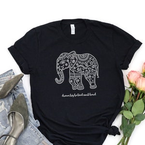 May include: A black short-sleeved t-shirt featuring a white graphic of an elephant decorated with intricate floral and geometric patterns. Below the elephant, the text reads "dream big be kind work hard" in a white cursive script font.