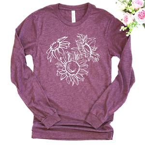 May include: A long-sleeved, burgundy-colored shirt featuring a white outline of three sunflowers. The floral design is centered on the front of the shirt. The shirt is made of a soft material and has a crew neck.