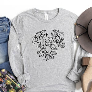 May include: A light gray long-sleeve shirt featuring a black line drawing of three sunflowers. The shirt is paired with blue jeans, a floral wallet, a gray hat, and tan ankle boots. The shirt is made of a soft, comfortable material.