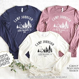 May include: Three long-sleeve shirts in navy blue, dusty rose, and cream. Each shirt features a white graphic with the text "CAMP JOHNSON" above a mountain and tree design, with "Sierra Meadows, CA 2024" below. The shirts are for camping.