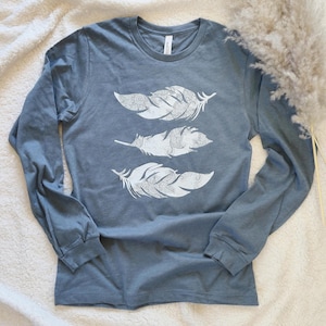 May include: A long-sleeved, crew neck shirt in a muted blue-grey hue. The shirt displays three white feather designs, each with detailed patterns. The feathers are a key feature, adding a bohemian style to the garment. The shirt is a casual wear item.