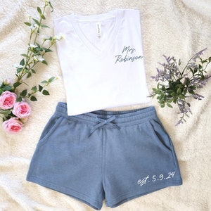 May include: A white V-neck t-shirt with "Mrs. Robinson" printed on the front, paired with blue shorts. The shorts have a drawstring and the text "est. 5.9.24". The set is displayed on a soft, textured surface.