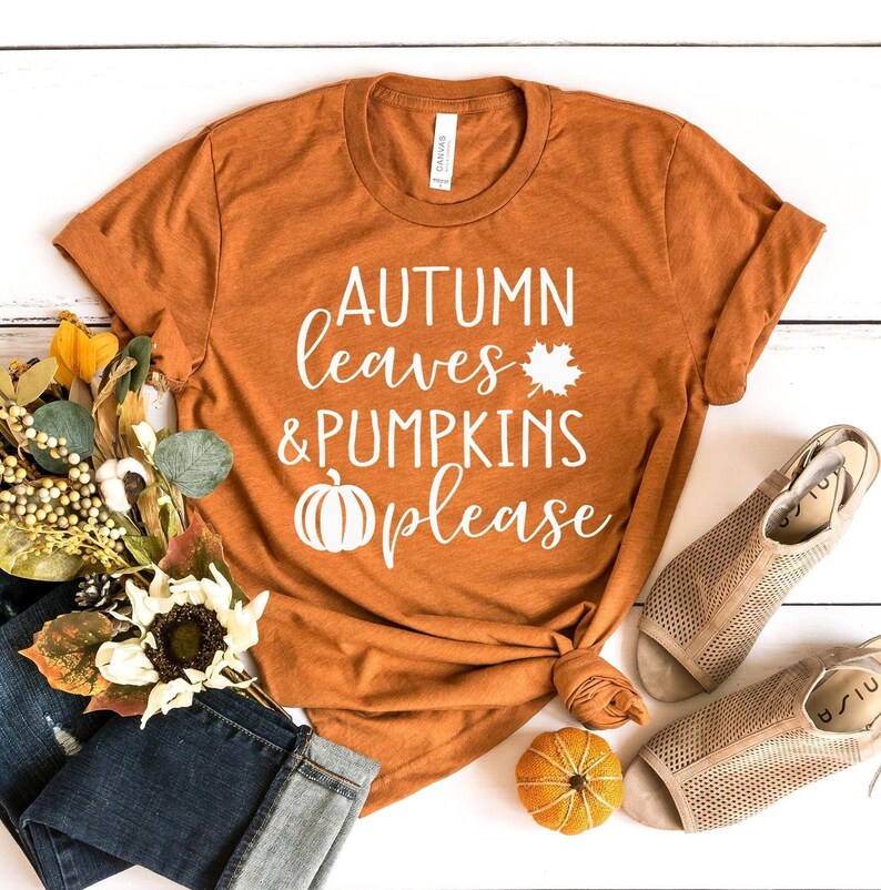 May include: An orange t-shirt with the text "AUTUMN leaves & PUMPKINS please" in white. The shirt is styled with a knot at the bottom. The image includes a pair of jeans, sandals, and fall-themed decorative items.