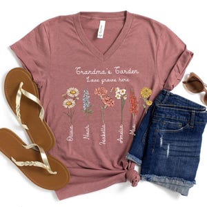 May include: A mauve V-neck t-shirt with the text "Grandma's Garden, Love grows here" and floral illustrations. The shirt is paired with denim shorts, sunglasses, and gold-strap sandals.