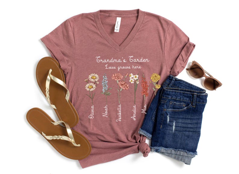 May include: A mauve V-neck t-shirt with the text "Grandma's Garden, Love grows here" and floral illustrations. The shirt is paired with denim shorts, sunglasses, and gold-strap sandals.