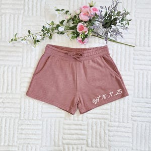 Op de afbeelding: Stofroze shorts met een trekkoord in de taille en zakken. De shorts hebben de tekst "est. 10.11.25" in witte letters op de rechterpijp. Een boeket roze en paarse bloemen ligt erboven.