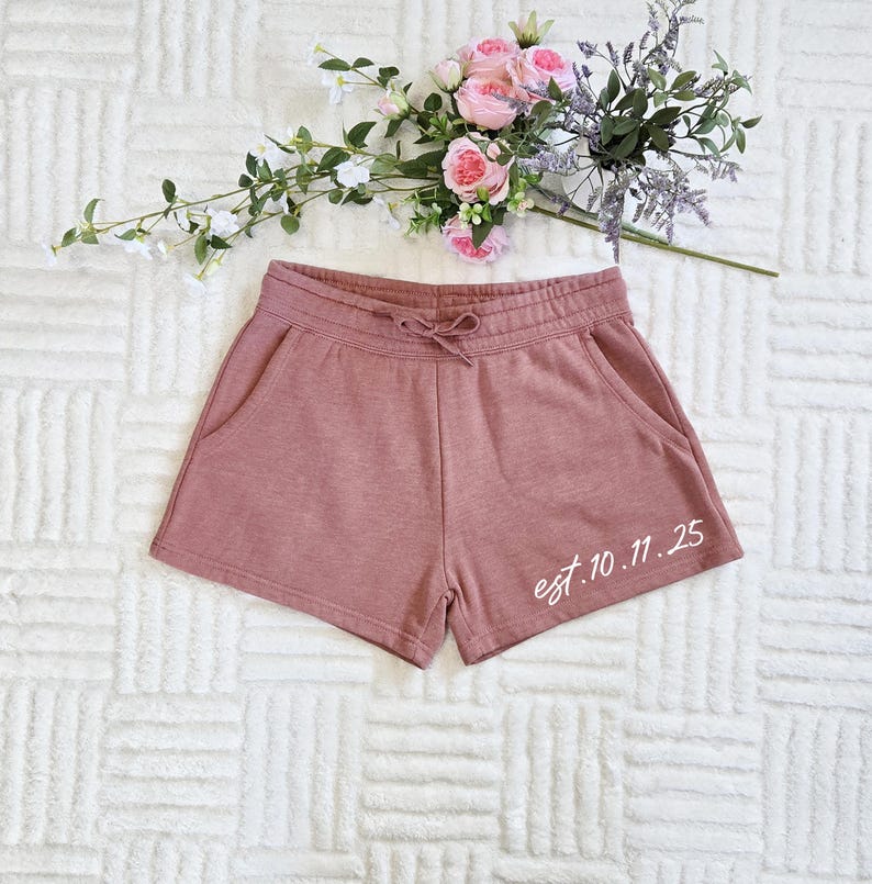 Op de afbeelding: Stofroze shorts met een trekkoord in de taille en zakken. De shorts hebben de tekst "est. 10.11.25" in witte letters op de rechterpijp. Een boeket roze en paarse bloemen ligt erboven.