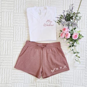 Op de afbeelding: Een witte V-hals T-shirt met roze "Mrs. Walker" tekst, gecombineerd met mauve shorts. De shorts hebben een taille met trekkoord en de tekst "est. 10.11.25". Een bloemstuk met roze rozen en paarse accenten maakt het ensemble compleet.