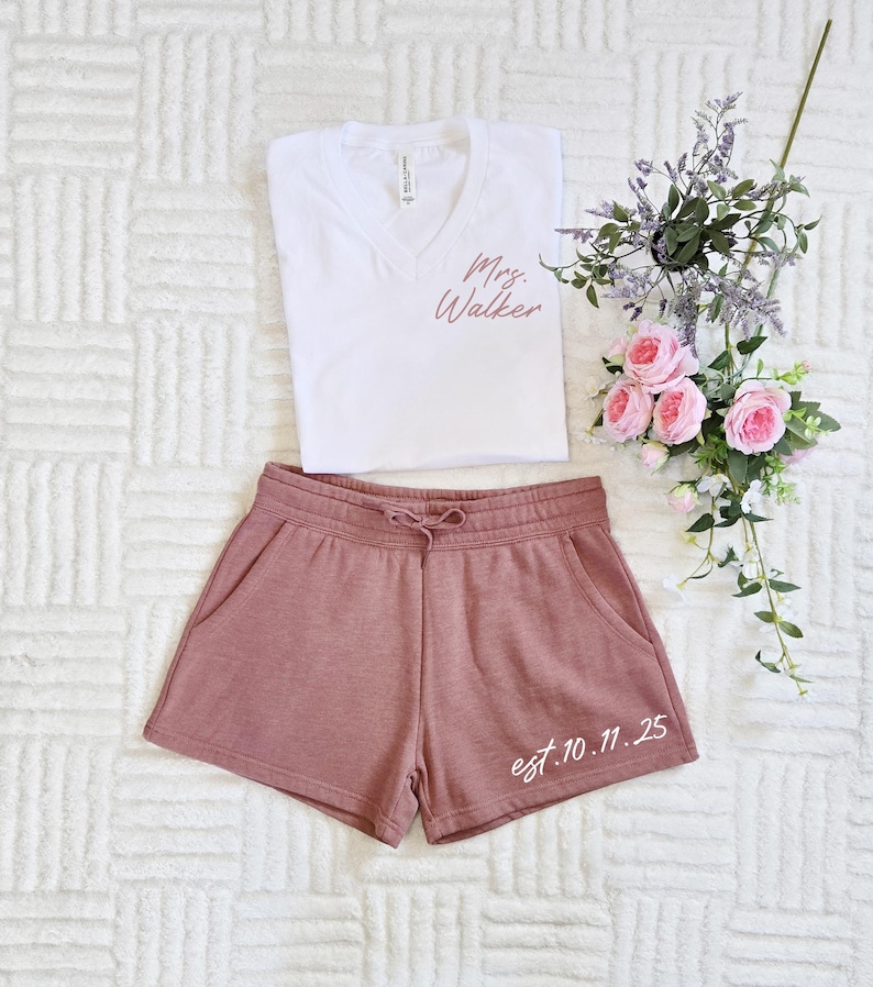 Op de afbeelding: Een witte V-hals T-shirt met roze "Mrs. Walker" tekst, gecombineerd met mauve shorts. De shorts hebben een taille met trekkoord en de tekst "est. 10.11.25". Een bloemstuk met roze rozen en paarse accenten maakt het ensemble compleet.