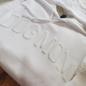 May include: White hoodie with the text "DOG MOM" in raised, light-colored lettering. The hoodie features a drawstring hood and a kangaroo pocket. The fabric appears to be a soft, comfortable material.