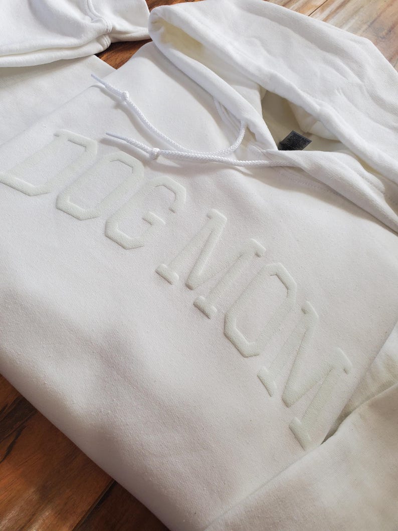 May include: White hoodie with the text "DOG MOM" in raised, light-colored lettering. The hoodie features a drawstring hood and a kangaroo pocket. The fabric appears to be a soft, comfortable material.