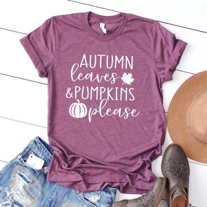 May include: A maroon t-shirt with the text "AUTUMN leaves & PUMPKINS please" in white. The design includes a maple leaf and a pumpkin graphic. The shirt is paired with denim shorts, a brown hat, and brown leather boots.
