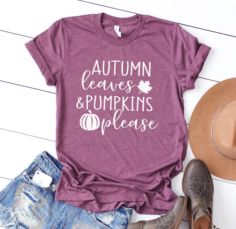 May include: A maroon t-shirt with the text "AUTUMN leaves & PUMPKINS please" in white. The design includes a maple leaf and a pumpkin graphic. The shirt is paired with denim shorts, a brown hat, and brown leather boots.