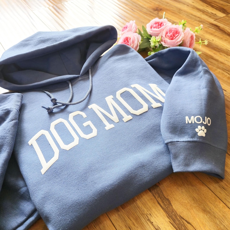 May include: A light blue hoodie with the words "DOG MOM" in white block letters across the front. The sleeve has the name "MOJO" and a paw print. The hoodie has a drawstring and hood.