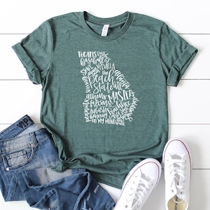 May include: A green t-shirt with the outline of the state of Georgia filled with white text. The text includes words like "Pecans", "Baseball", "Athens", and "Masters". The shirt is paired with blue jeans and white sneakers.