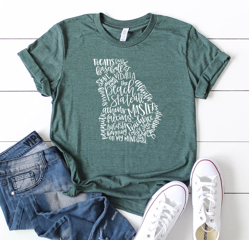 May include: A green t-shirt with the outline of the state of Georgia filled with white text. The text includes words like "Pecans", "Baseball", "Athens", and "Masters". The shirt is paired with blue jeans and white sneakers.