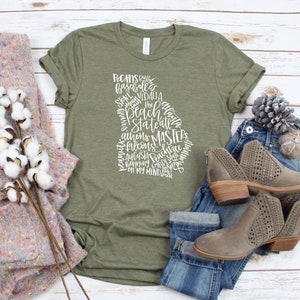 May include: An olive green t-shirt with a white graphic in the shape of the state of Georgia. The graphic includes the words "Pecans", "Peach State", "Atlanta", and other Georgia-related terms. The shirt is paired with jeans and brown ankle boots.
