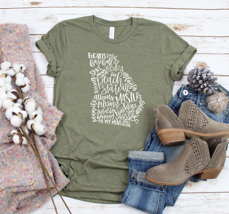 May include: An olive green t-shirt with a white graphic in the shape of the state of Georgia. The graphic includes the words "Pecans", "Peach State", "Atlanta", and other Georgia-related terms. The shirt is paired with jeans and brown ankle boots.