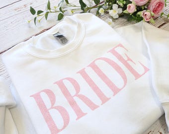 May include: A white crew neck sweatshirt with the word "BRIDE" printed in large, pink, glittery letters. The sweatshirt is folded and placed on a light wood surface, with pink roses and greenery in the background.
