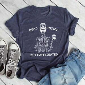 May include: A navy blue t-shirt with a white graphic of a skeleton wearing sunglasses and holding a coffee cup. The text on the shirt reads "DEAD INSIDE BUT CAFFEINATED". Gray sneakers and blue jeans are next to the shirt.