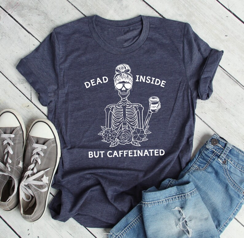 May include: A navy blue t-shirt with a white graphic of a skeleton wearing sunglasses and holding a coffee cup. The text on the shirt reads "DEAD INSIDE BUT CAFFEINATED". Gray sneakers and blue jeans are next to the shirt.