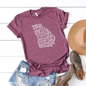 May include: Burgundy t-shirt with a white graphic in the shape of the state of Georgia, filled with words like "Pecans," "Braves," and "Masters." The shirt is paired with denim shorts, a brown hat, and gray shoes.