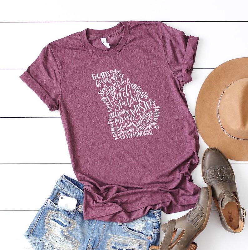 May include: Burgundy t-shirt with a white graphic in the shape of the state of Georgia, filled with words like "Pecans," "Braves," and "Masters." The shirt is paired with denim shorts, a brown hat, and gray shoes.