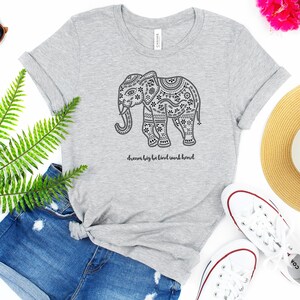 May include: A heather gray short-sleeve t-shirt featuring a black graphic of an elephant decorated with intricate floral and geometric patterns. Below the elephant, the text reads, dream big be kind work hard. The shirt has a crew neckline.