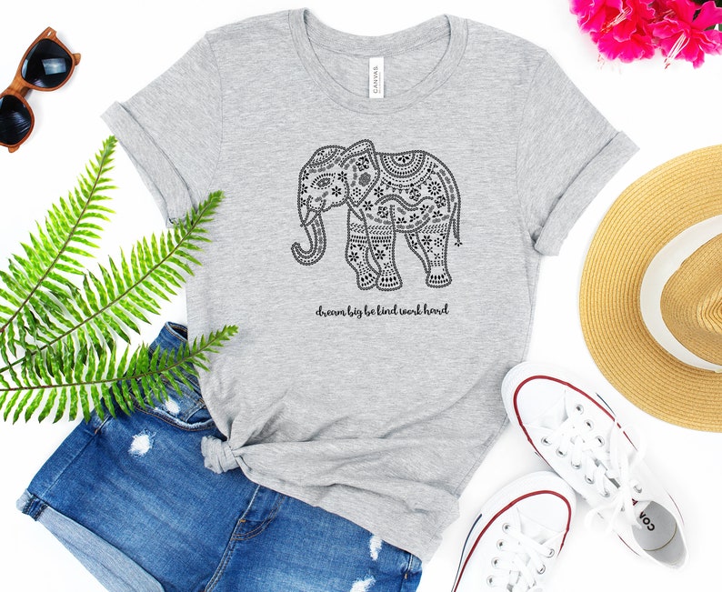 May include: A heather gray short-sleeve t-shirt featuring a black graphic of an elephant decorated with intricate floral and geometric patterns. Below the elephant, the text reads, dream big be kind work hard. The shirt has a crew neckline.