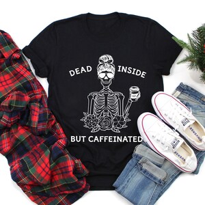 May include: Black t-shirt with a white skeleton graphic holding a coffee cup, with the text "DEAD INSIDE BUT CAFFEINATED." The shirt is paired with blue jeans, white sneakers, and a red and black plaid shirt.
