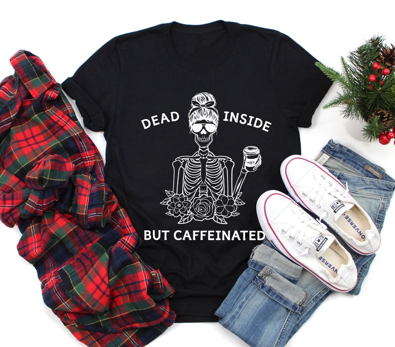 May include: Black t-shirt with a white skeleton graphic holding a coffee cup, with the text "DEAD INSIDE BUT CAFFEINATED." The shirt is paired with blue jeans, white sneakers, and a red and black plaid shirt.