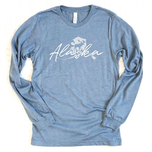 May include: A light blue long-sleeve shirt with the word "Alaska" in white script, along with a white graphic of the state's outline and a moose. The shirt has a crew neck and long sleeves with ribbed cuffs.