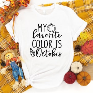 May include: White t-shirt with the text "MY favorite COLOR IS October" in black script. A pumpkin graphic is above the word "October". The shirt is surrounded by fall-themed decorations, including pumpkins and autumn leaves.
