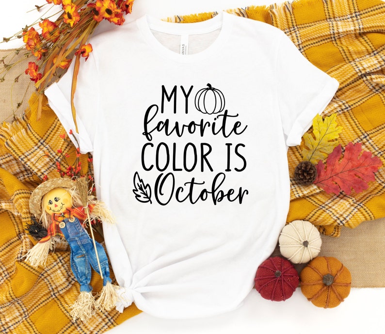 May include: White t-shirt with the text "MY favorite COLOR IS October" in black script. A pumpkin graphic is above the word "October". The shirt is surrounded by fall-themed decorations, including pumpkins and autumn leaves.