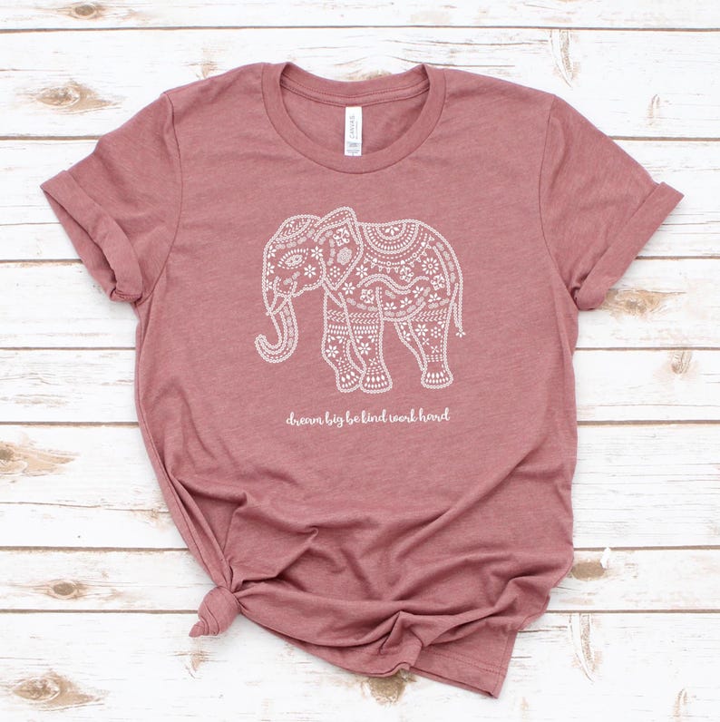 May include: A mauve short-sleeved t-shirt featuring a white graphic of an elephant decorated with intricate floral and geometric patterns. Below the elephant, the text "dream big be kind work hard" is printed in a white, cursive script font.