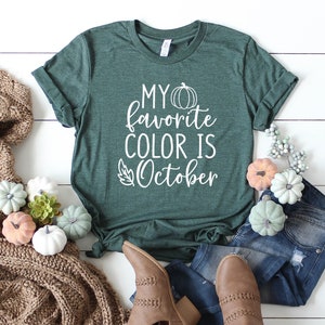 May include: A dark green t-shirt with the text "MY favorite COLOR IS October" in white. The shirt is styled with blue jeans, brown boots, and decorative pumpkins in various colors. A cozy brown knit scarf is also included.
