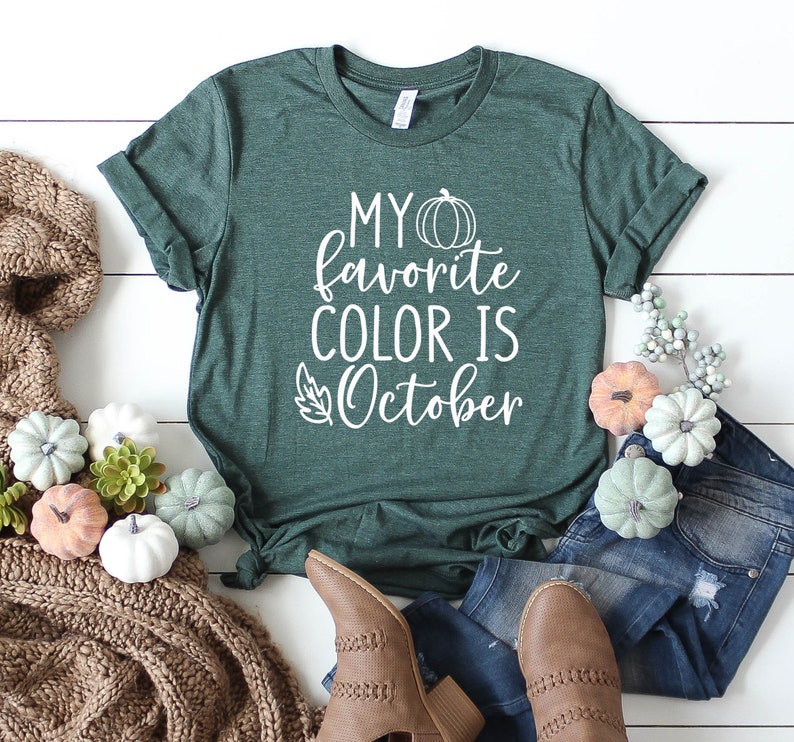 May include: A dark green t-shirt with the text "MY favorite COLOR IS October" in white. The shirt is styled with blue jeans, brown boots, and decorative pumpkins in various colors. A cozy brown knit scarf is also included.