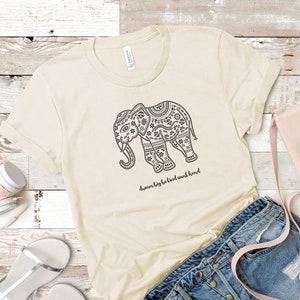 May include: A cream-colored short-sleeve t-shirt featuring a black graphic of an elephant decorated with intricate patterns. Below the elephant, the text reads "dream big be kind work hard" in a cursive black font. The shirt has a crew neckline.