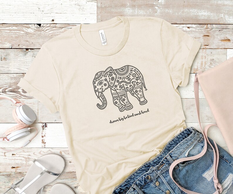 May include: A cream-colored short-sleeve t-shirt featuring a black graphic of an elephant decorated with intricate patterns. Below the elephant, the text reads "dream big be kind work hard" in a cursive black font. The shirt has a crew neckline.
