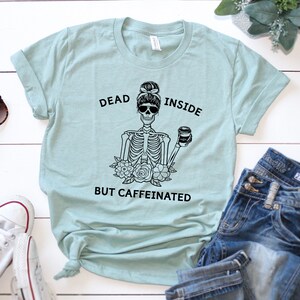 May include: A light blue t-shirt with the text "DEAD INSIDE BUT CAFFEINATED" and a black graphic of a skeleton with sunglasses holding a coffee cup. The shirt is paired with blue jeans and white sneakers.