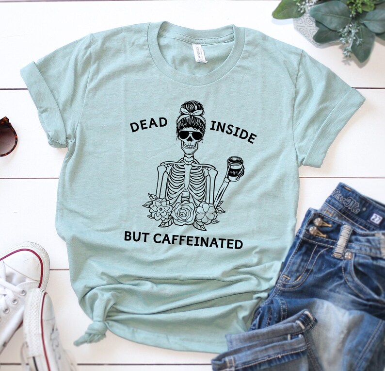May include: A light blue t-shirt with the text "DEAD INSIDE BUT CAFFEINATED" and a black graphic of a skeleton with sunglasses holding a coffee cup. The shirt is paired with blue jeans and white sneakers.