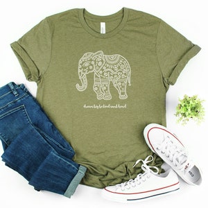 May include: An olive green short-sleeve t-shirt featuring a white graphic of an elephant filled with intricate floral patterns and the text "dream big be kind work hard" printed below the elephant, paired with blue denim jeans and white sneakers.